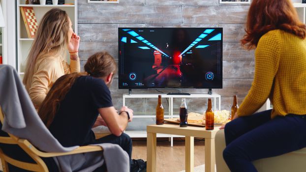 Back view of blonde woman playing shooter games while her friends are eating pizza. Playing games with wireless controller.