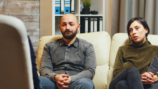 Young man breathing heavily sitting on couch with his wife at couple therapy talking about their relationship.