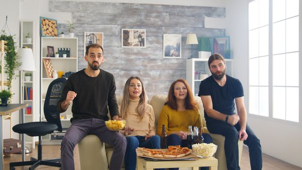 Revealing shot of close friends watching a football championship on tv sitting on couch eating pizza and drinking beer.