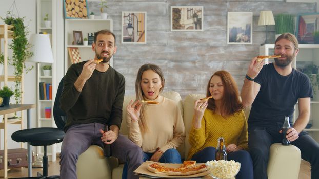 Man changing channels on tv using remote control while a tv show with his friends, drinking beer and eating pizza.