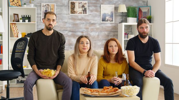 Young man clapping while watching a movie with his friends in living room eating pizza and drinking beer.