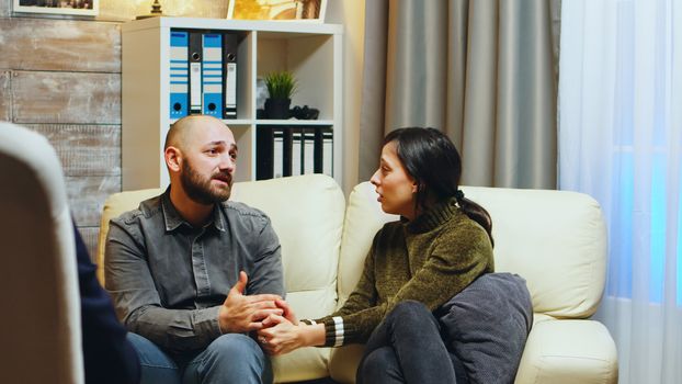 Sad girlfriend holding husband hands sitting on couch at couple therapy. Therapist giving guidance to stressed couple.