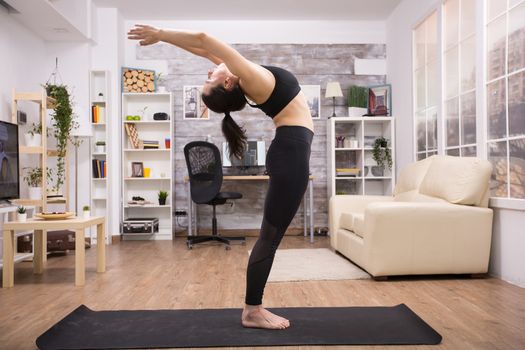 Young yoga practitioner standing doing backward stretching sitting on mat in home.