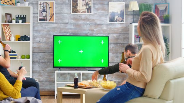 Back view of friends drinking beer and watching sports on tv with green screen in living room.