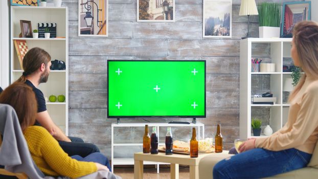Back view of friends drinking beer and watching sports on tv with green screen in living room.