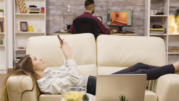 Beautiful young girl lying on the couch using her phone. Boyfriend in the background.