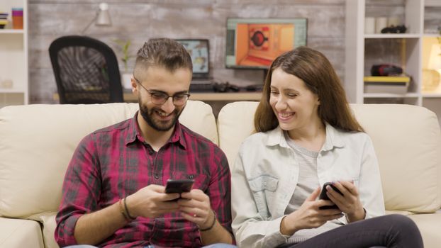 Couple laughing together while browsing on their phones. Couple sitting on couch.