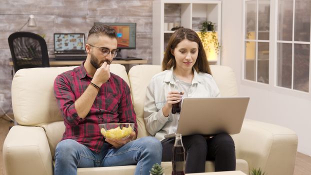 Happy couple on the couch eating chips and looking at a laptop