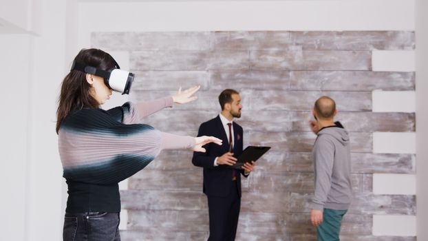 Female designer with virtual reality goggles in empty apartment while real estate agent is talking with the homeowner in the background.