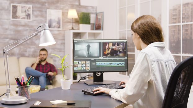 Side view of video editor working on a project from home. Boyfriend in the background is talking on the phone.