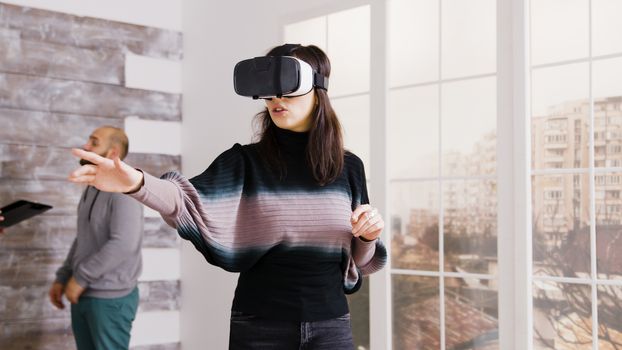 Female architect using virtual reality goggles in empty apartment and real estate agent talking with client in the background.