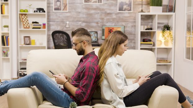 Unhappy couple sitting back to back on the couch. Couple ingoring each other using phones.