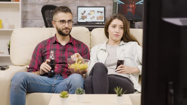Beautiful young couple sitting on the couch enjoying chips and soda while watching tv at night.