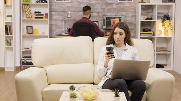 Girl using her phone and laptop while sitting on couch. Boyfriend in the background.