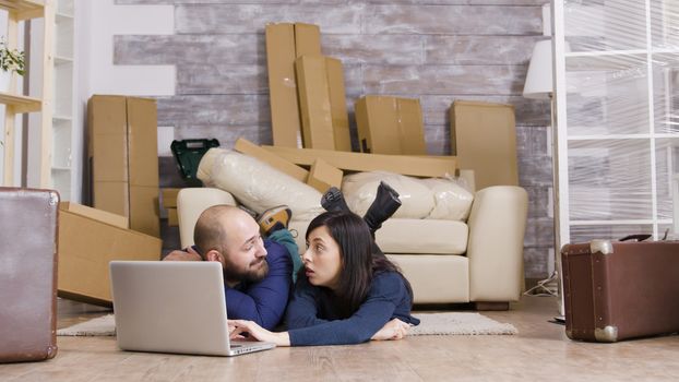 Couple laying down on the floor of their new apartment doing online shopping on laptop. Zoom in shot.