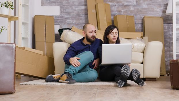 Couple sitting on the floor of new apartment and using laptop for online shopping.