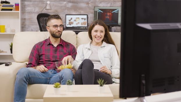 Charming young couple sitting on the couch and watching tv while enjoying their chips.