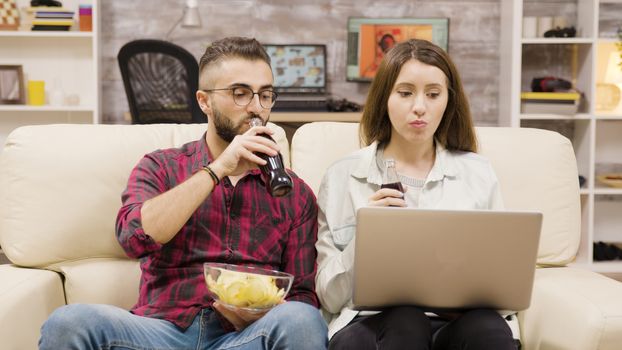 Couple drinking soda and eating chips while browsing on laptop. Couple relaxing on the couch.