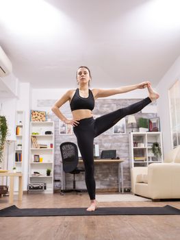 Brunette adult woman doing a standing leg yoga pose in living room.