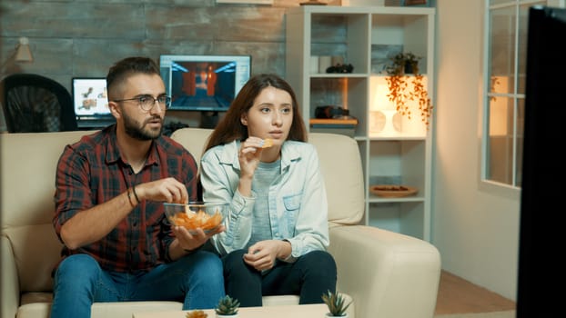 Young couple sitting on the couch and watching television enjoying their chips. Couple looking tense on television.