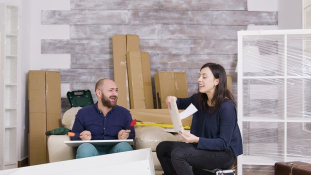 Young couple laughing while assembling furniture in their apartment. Girl reading instructions.