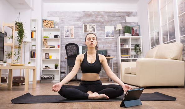 Young woman in peaceful state of mind while doing yoga with eyes closed and tablet computer in front of her.
