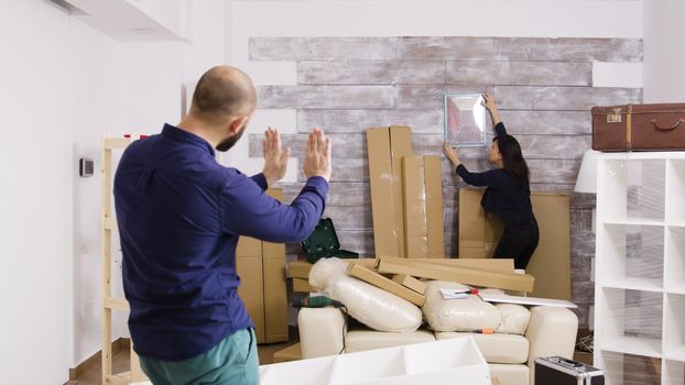 Caucasian boyfriend telling his girlfriend how to hang picture in the wall of their new apartment. Couple decorating home wall.
