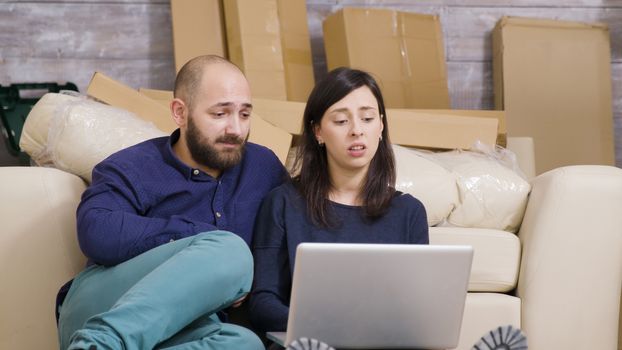 Couple in love just moved in their new apartment and making online shopping on laptop. Cardboard boxes in the background.