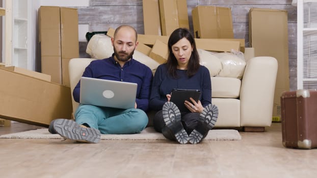 Couple in their new flat sitting on the floor shopping online for furniture using laptop and tablet.