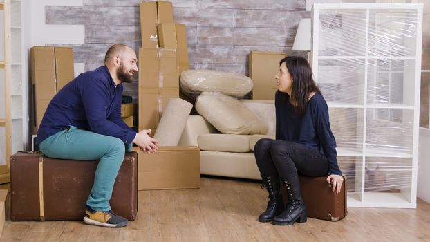 Couple talking and sitting on suitcases after carrying boxes. Cardboard boxes in the background.