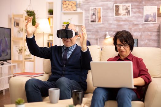 Old elderly retired man using VR virtual reality headset in their cozy apartment.