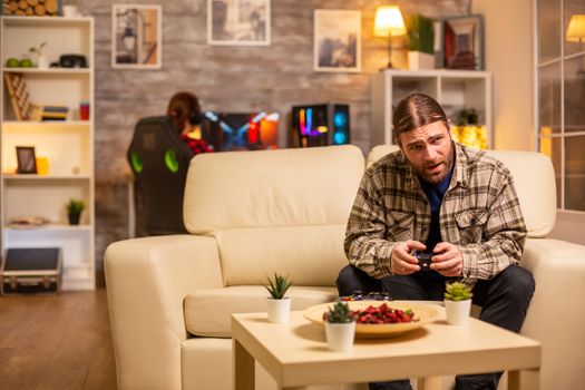 Male gamer playing video games on a console in the living room while sitting on the sofa