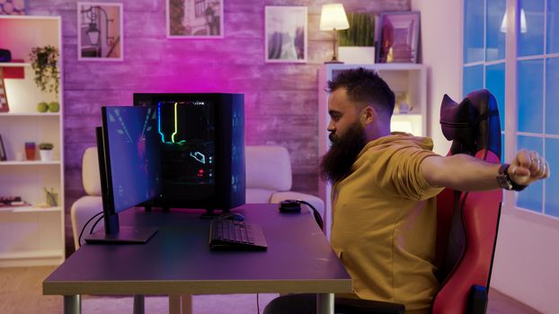 Bearded man sitting on gaming chair stretching while playing video games on pc. Colorful neon light in the room