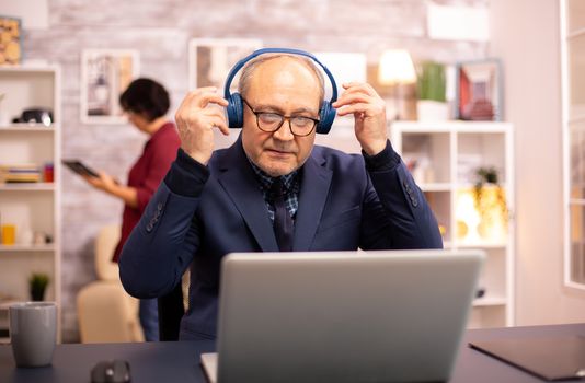 Elderly man in his 60s with headphones on his head listening to music and working on a modern laptop