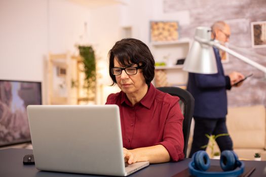 Old woman using a modern computer in her living room while her husband walks in the background