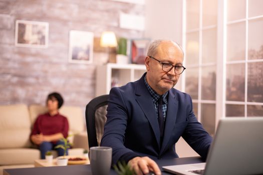 Old man in his 60s working on a laptop in cozy living room while his wife is in background