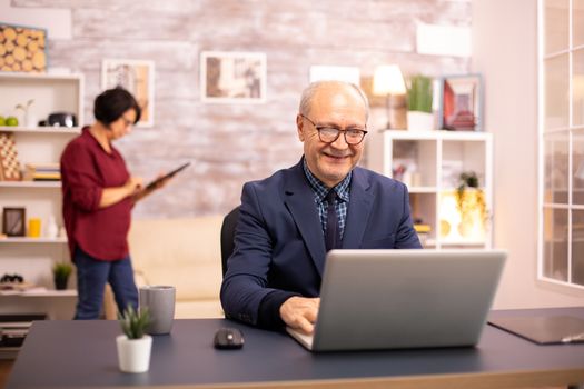 Successful elderly person in suit working on a laptop from home. His wife is in the background