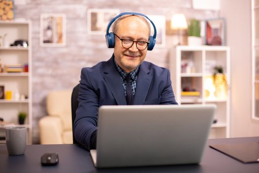 Elderly man in his 60s with headphones on his head listening to music and working on a modern laptop