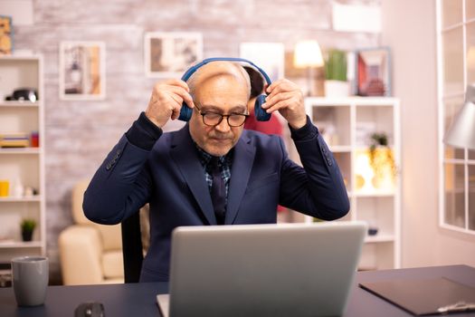 Elderly man in his 60s with headphones on his head listening to music and working on a modern laptop