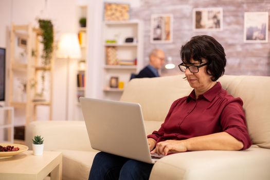 Old elderly woman on her sofa working on a modern laptop in her cozy living room. Her husband is in the background