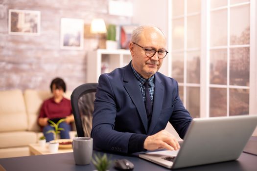 Old man in his 60s working on a laptop in cozy living room while his wife is in background