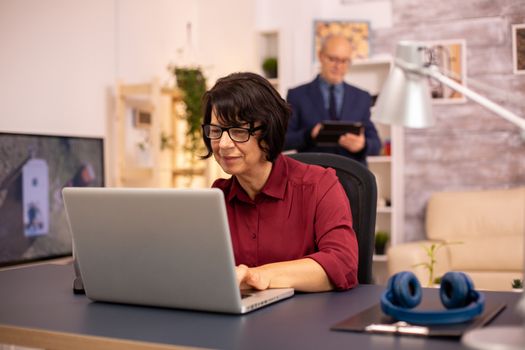 Old woman using a modern computer in her living room while her husband walks in the background