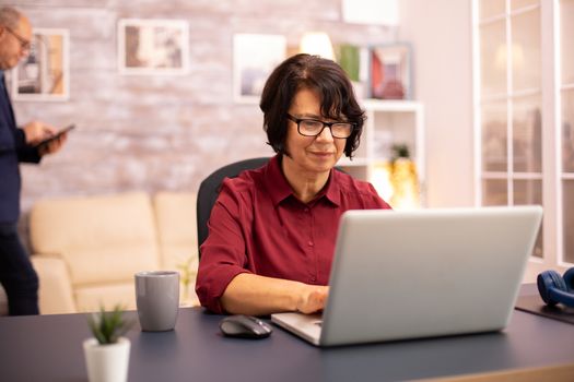 Old woman in her 60s using a modern laptop in her cozy house late in the evening