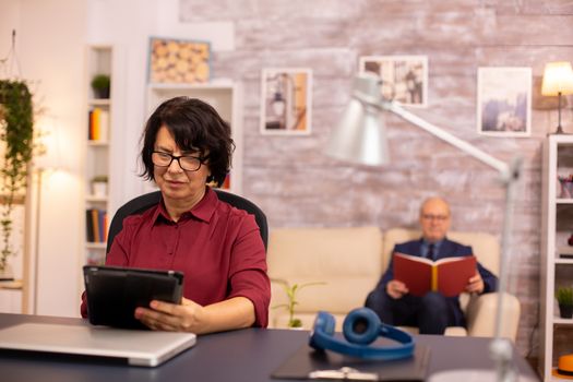 Old lady in her 60s using modern technolgy in cozy living space while her husband reads in the background