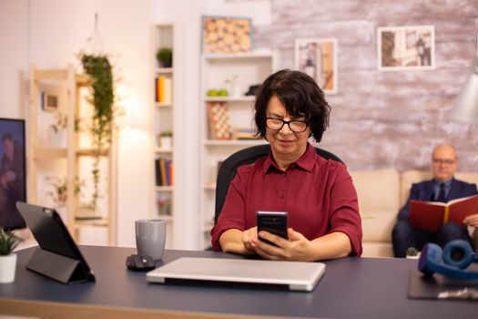 Old lady in her 60s using modern technolgy in cozy living space while her husband reads in the background