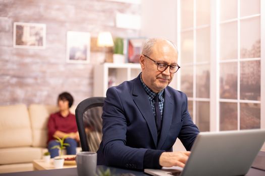 Old man in his 60s working on a laptop in cozy living room while his wife is in background