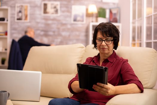 Old elderly woman sitting on the sofa and using a digital tablet PC in cozy living room.