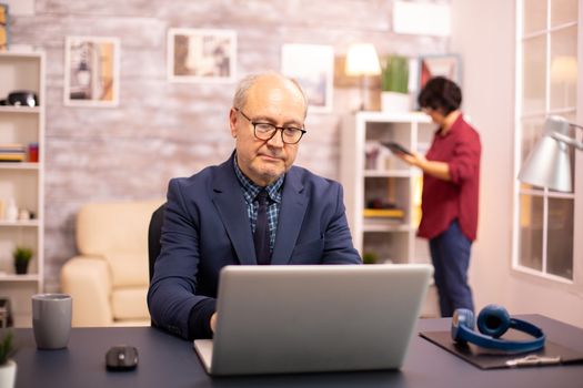 Successful elderly person in suit working on a laptop from home. His wife is in the background