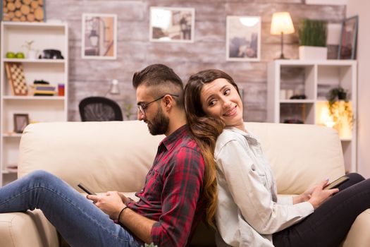 Girlfriend looking at her boyfriend while sitting back to back on sofa after and disagreement. Boyfriend ignoring girlfriend.