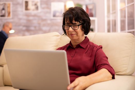 Old elderly woman on her sofa working on a modern laptop in her cozy living room. Her husband is in the background